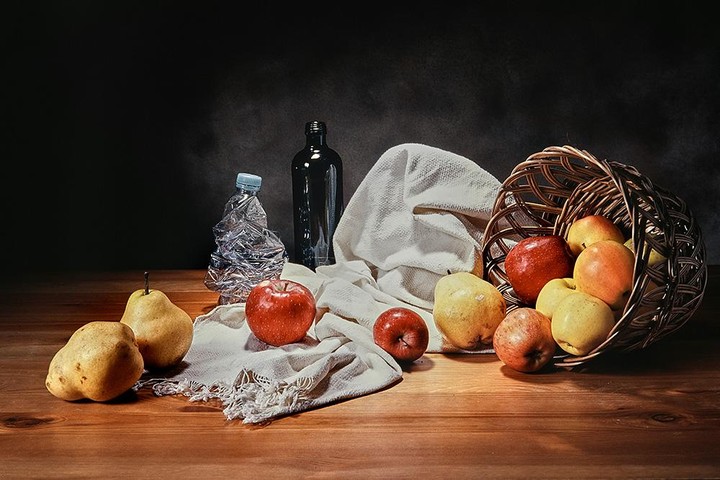 A still life arrangement features a wicker basket spilling apples and pears onto a wooden surface, alongside a crumpled plastic bottle, a glass bottle, and a white cloth.