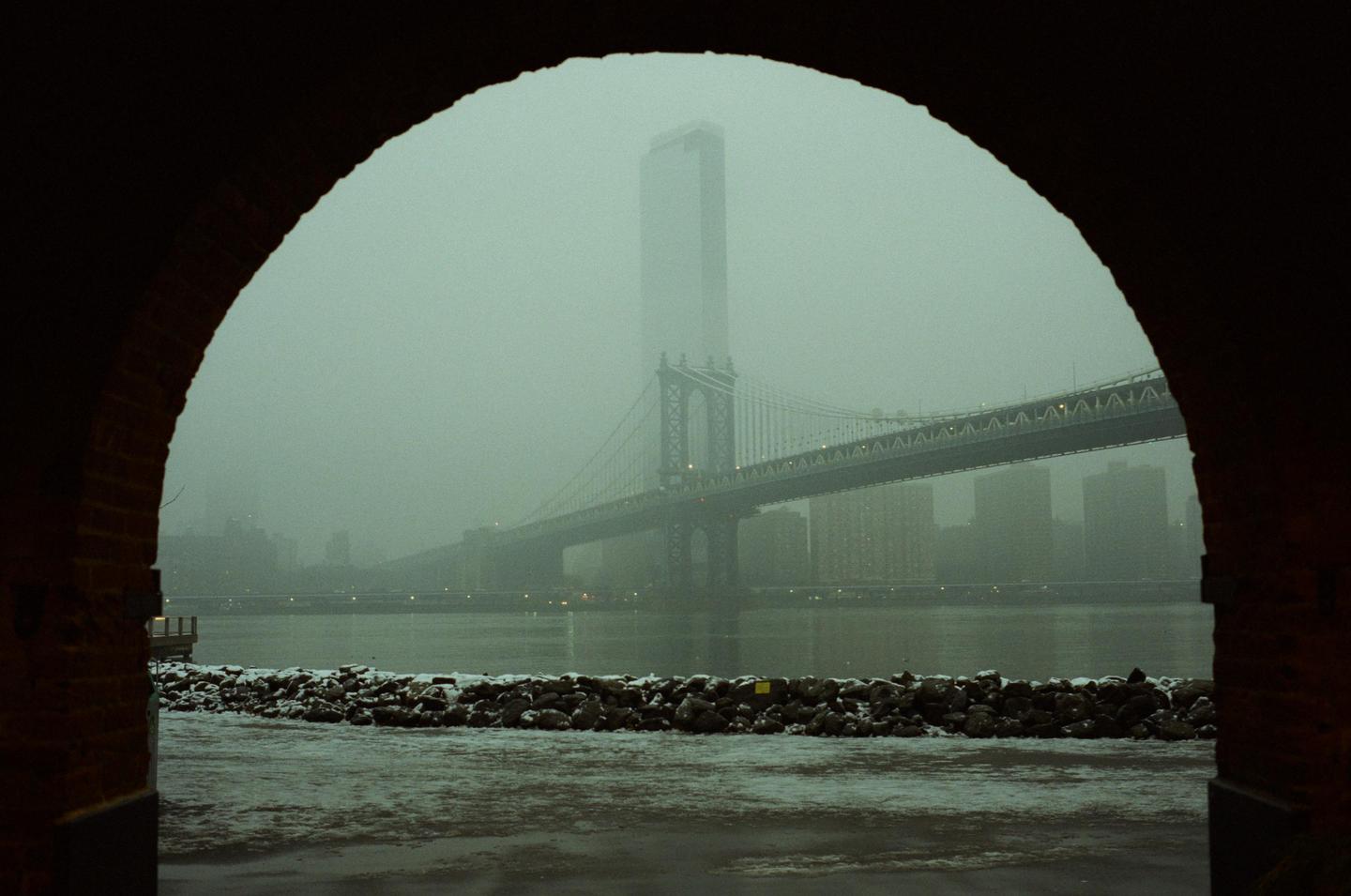 A view through an archway shows a bridge and a tall building across a river on a foggy day.