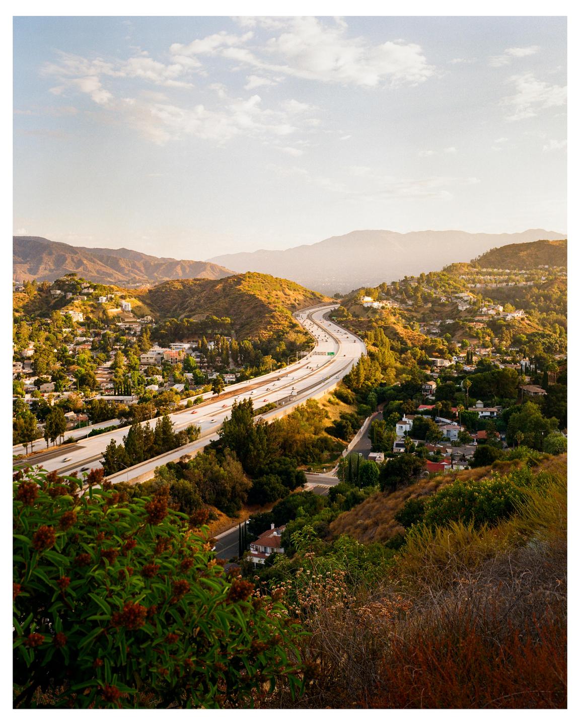 A freeway winds through a valley filled with houses and trees, with mountains in the distance under a hazy sky.