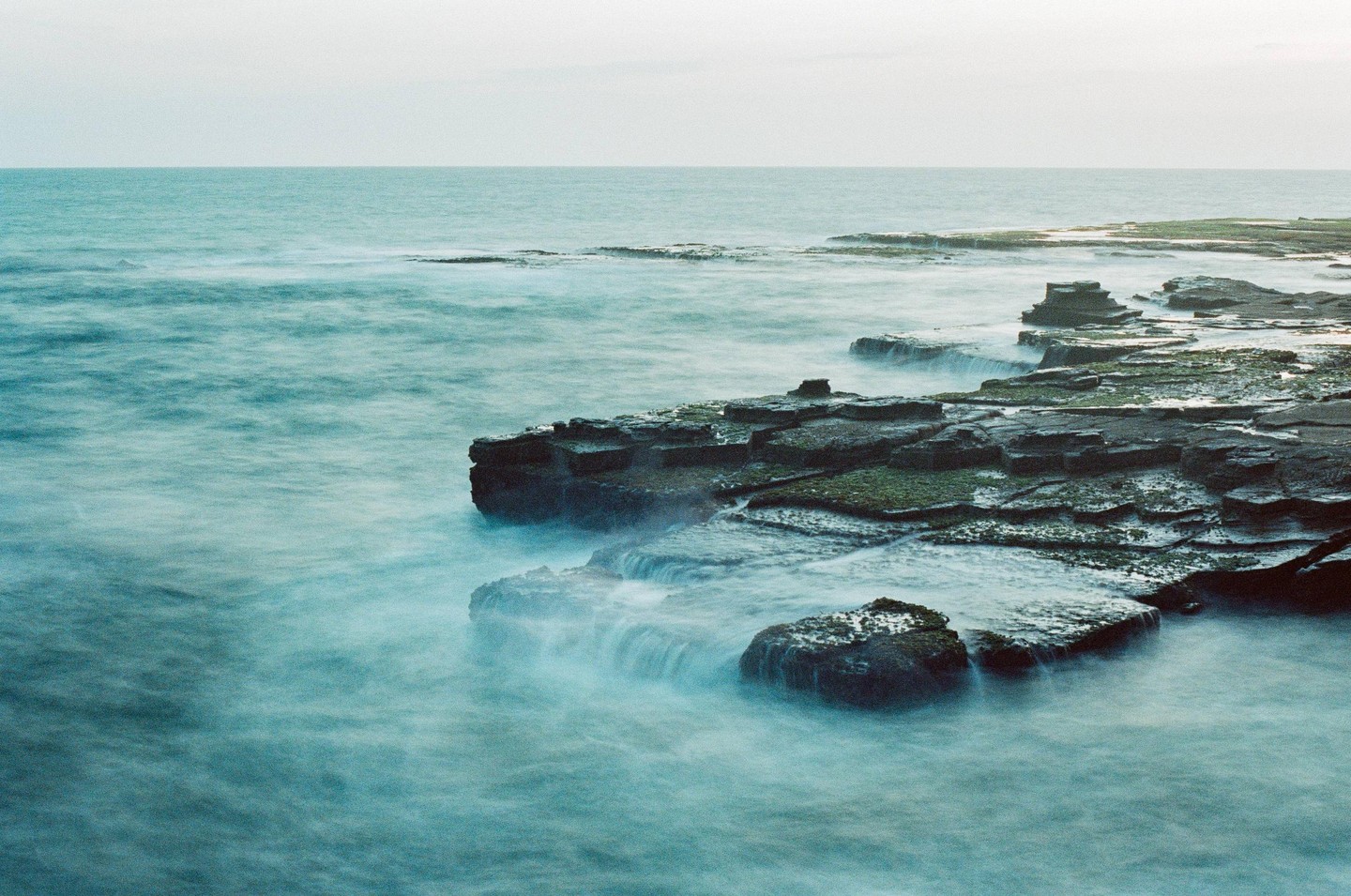 The ocean washes over a rocky coastline under a pale sky.