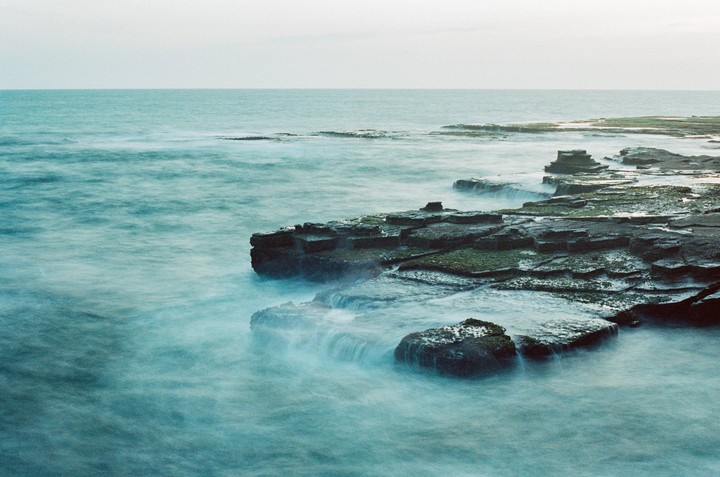 The ocean washes over a rocky coastline under a pale sky.