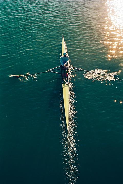 A person rows a single scull boat on a body of water with sun glare.