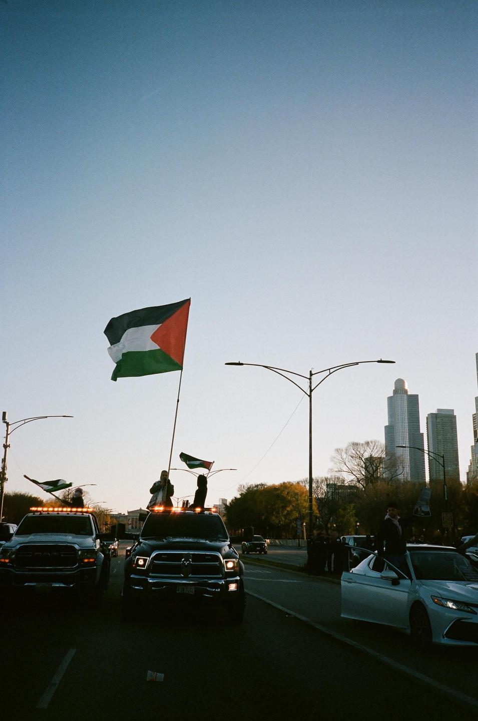 A large Palestinian flag waves from a truck on a city street with buildings in the background.