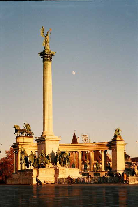 A tall column with a winged statue on top stands in a plaza with other statues and a building in the background under a clear sky with the moon visible.