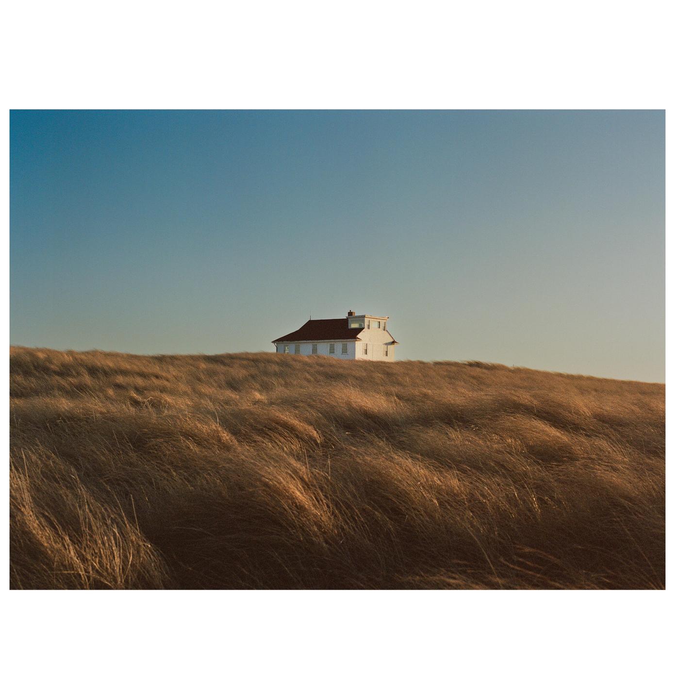 A white house with a red roof sits atop a hill covered in tall, dry grass under a clear sky.