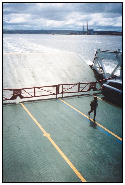 A child runs across the deck of a ferry with a body of water and industrial buildings in the background.