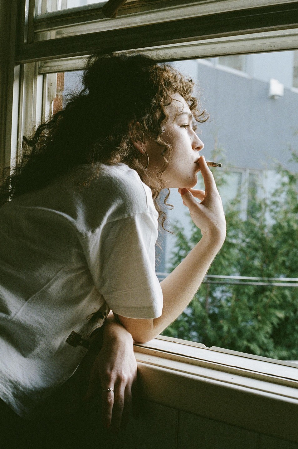 A woman with curly hair leans out a window, smoking a cigarette and looking into the distance.