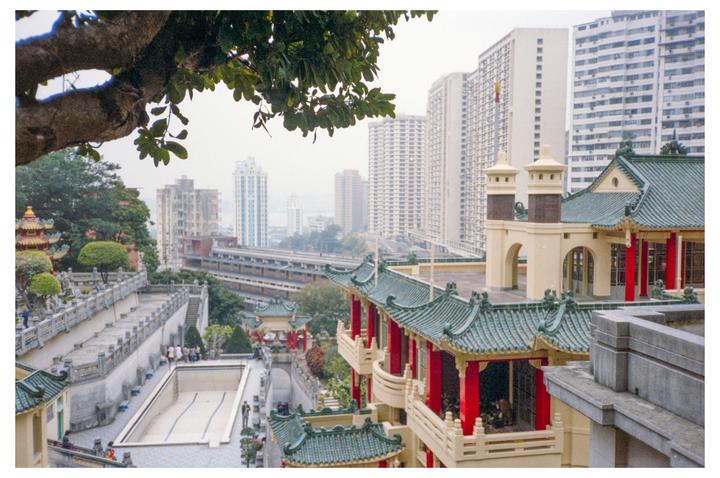 A traditional Chinese temple complex with green roofs and red pillars is nestled among modern high-rise buildings.