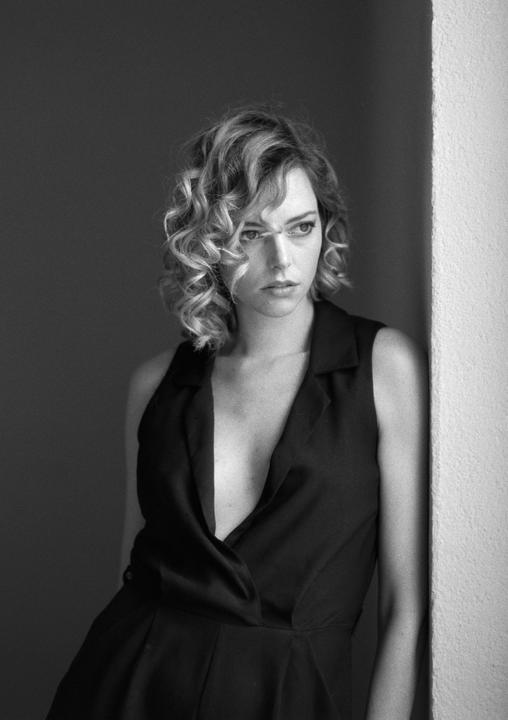 A woman with curly hair and a dark top leans against a wall.