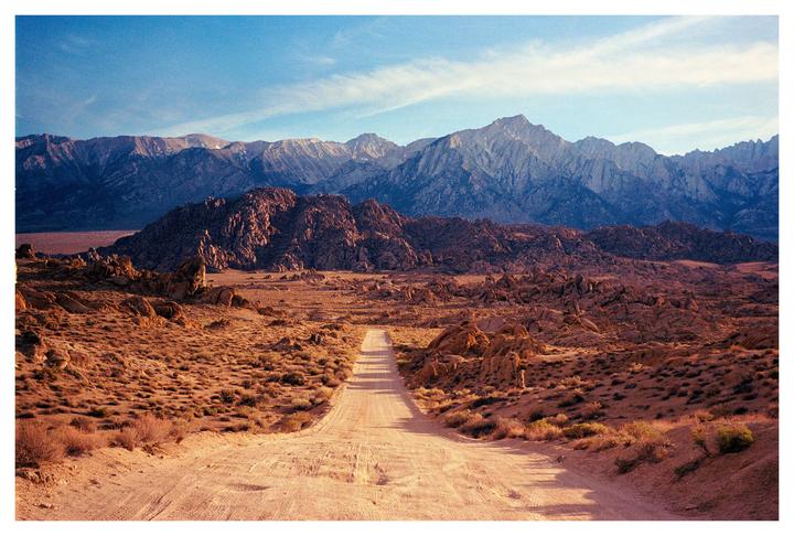 A dirt road leads through a desert landscape towards mountains under a blue sky.