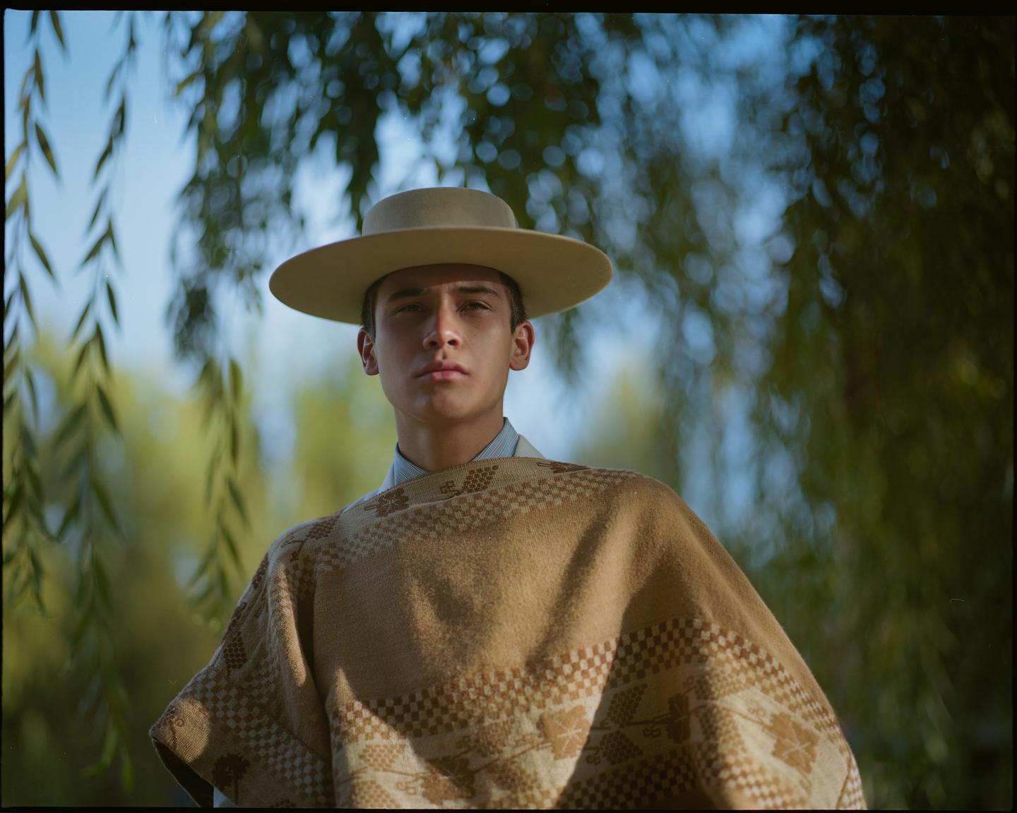 A young man wearing a poncho and a wide-brimmed hat stands outdoors with green foliage in the background.
