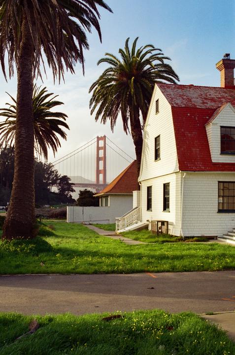 A white house with a red roof stands next to palm trees, with the Golden Gate Bridge visible in the background.