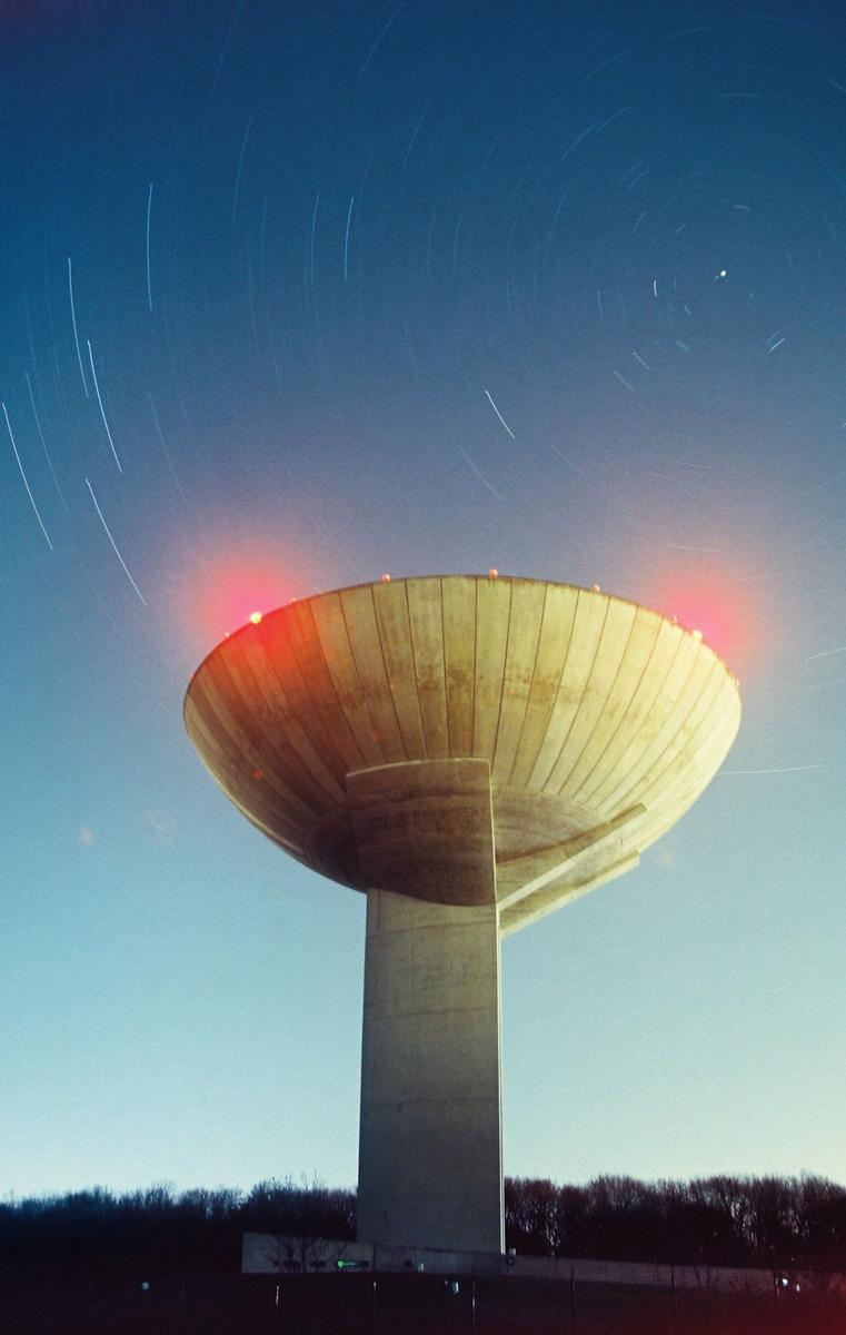 A concrete water tower with red lights on its rim stands against a blue sky with star trails.