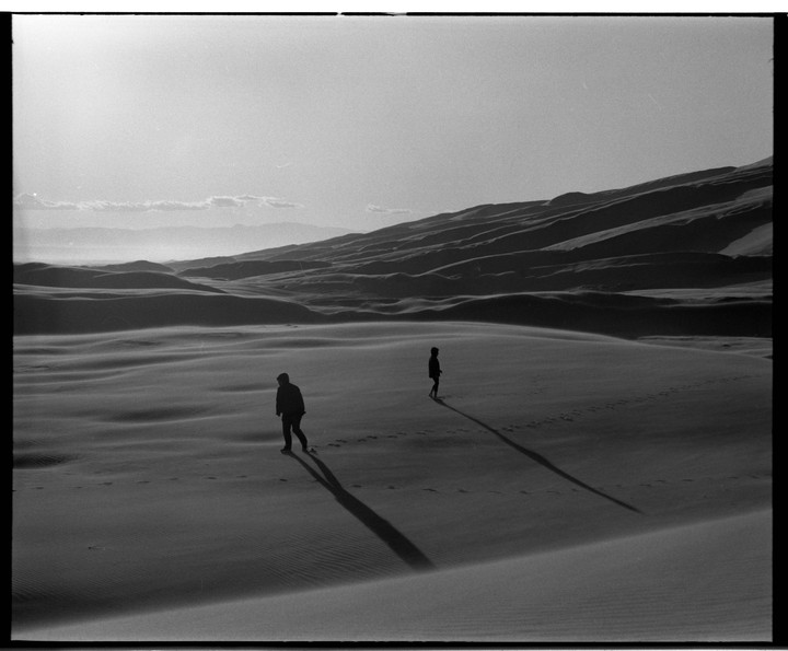 Two people walk across sand dunes, casting long shadows.