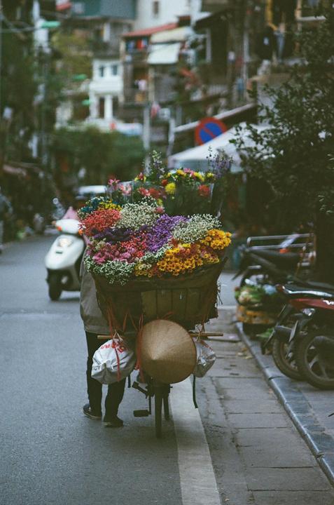 A person pushes a bicycle laden with a large basket of colorful flowers and a conical hat on a city street.