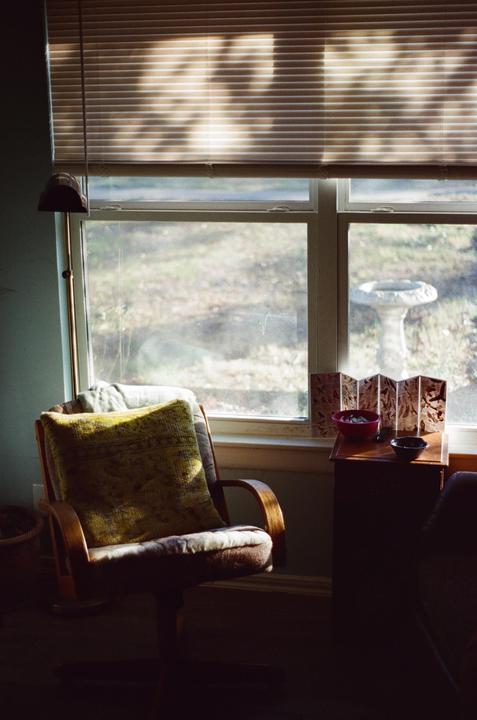 Sunlight streams through a window with blinds, illuminating a chair with a pillow and a small table with bowls and a decorative screen.