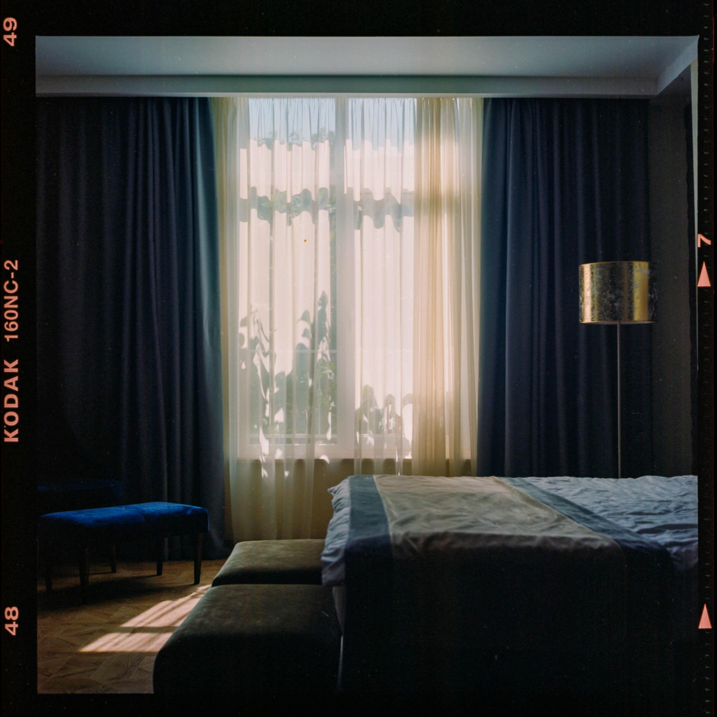 Sunlight streams through a window with sheer and dark curtains into a bedroom with a bed, benches, and a floor lamp.