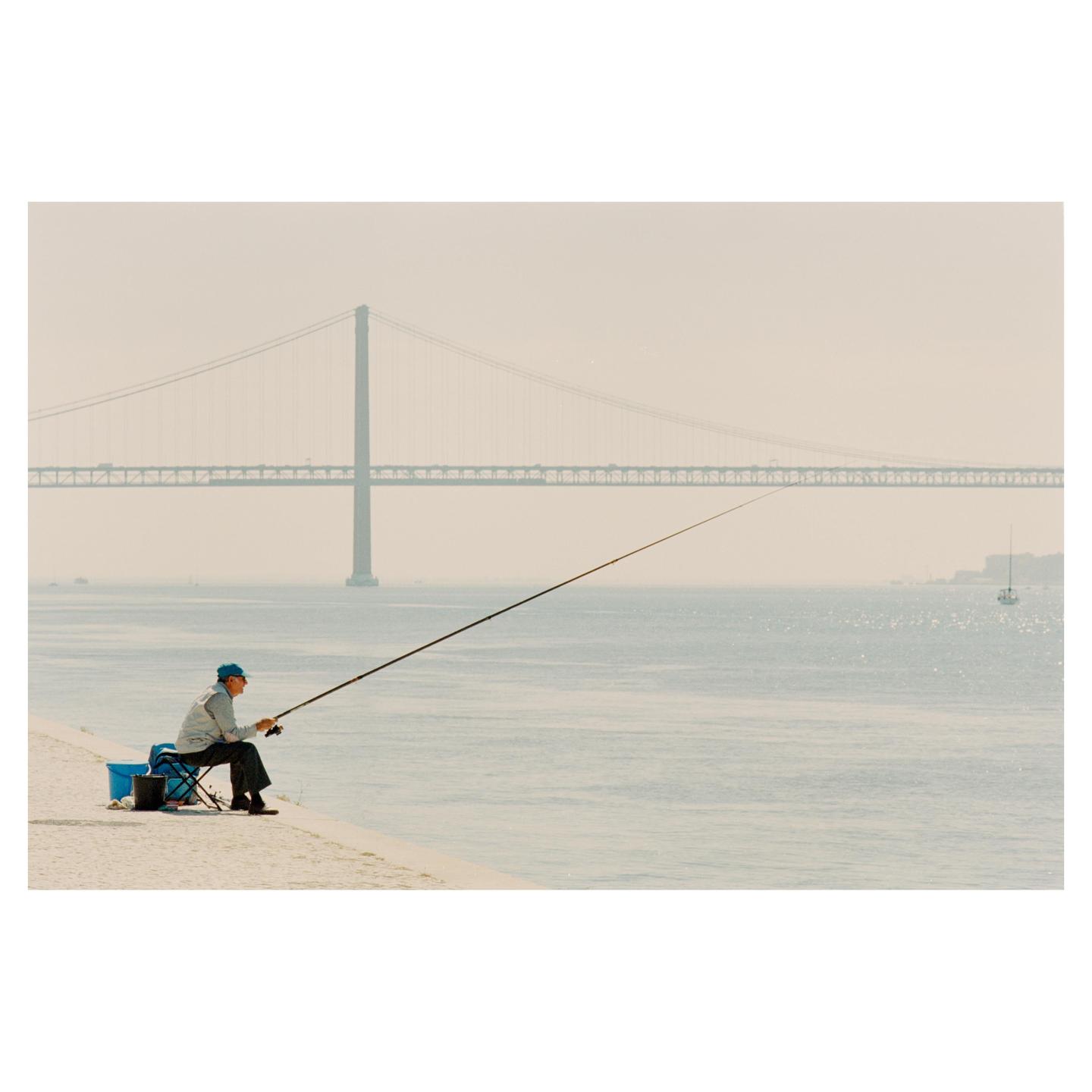 A man sits on a stool fishing in a body of water with a large suspension bridge in the background.