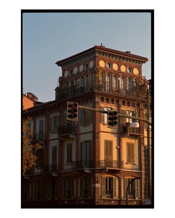 A brick building with balconies and a decorative top floor is illuminated by golden hour light, with traffic lights in the foreground.
