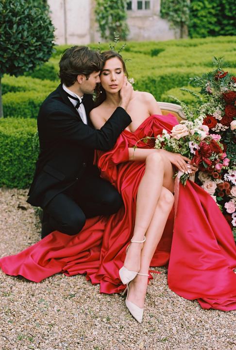 A man in a tuxedo kneels beside a woman in a red dress, who is seated on a couch with a bouquet of flowers next to her.