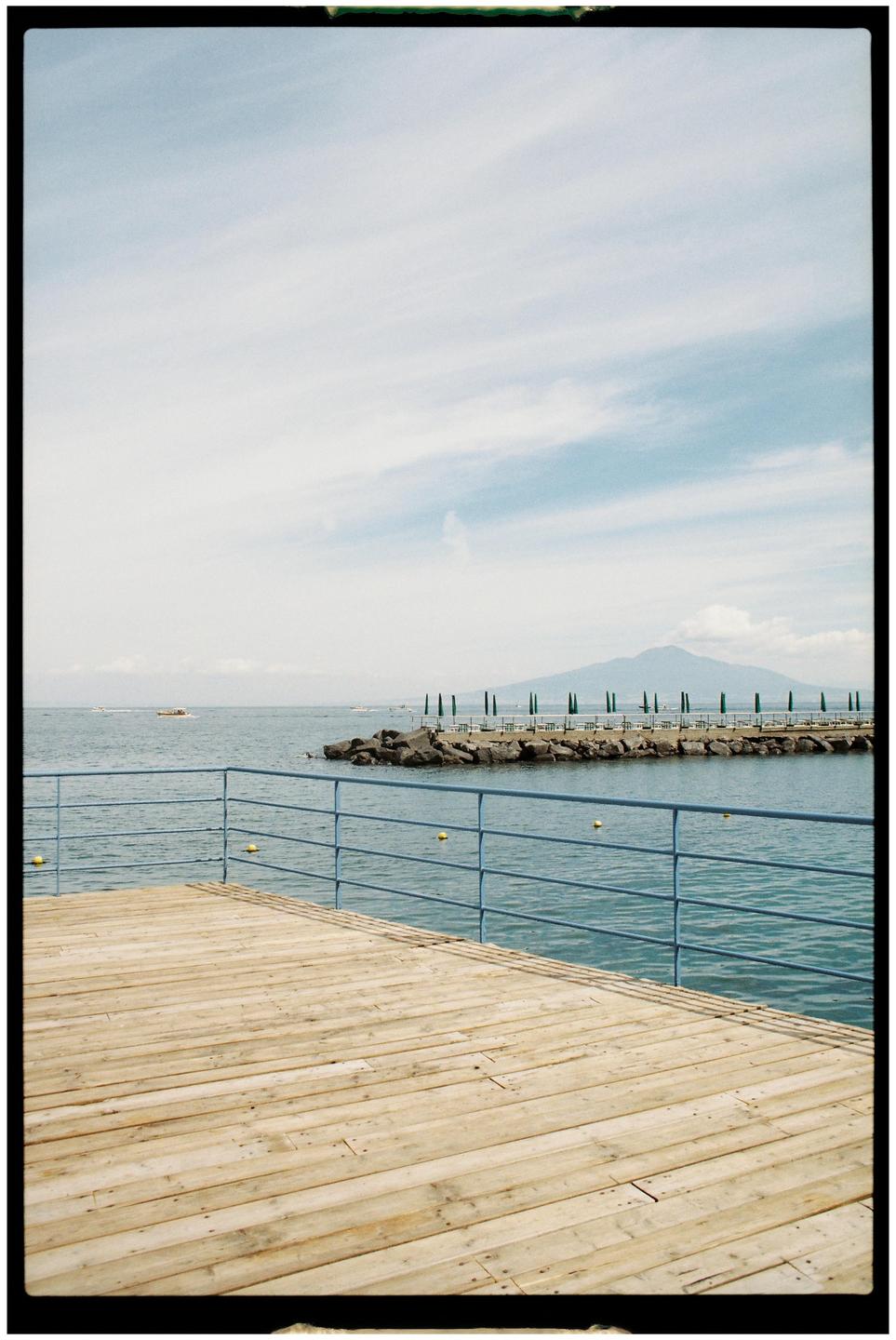 A wooden pier with a blue railing extends over the water towards a rocky breakwater lined with green beach umbrellas and a distant mountain under a cloudy sky.