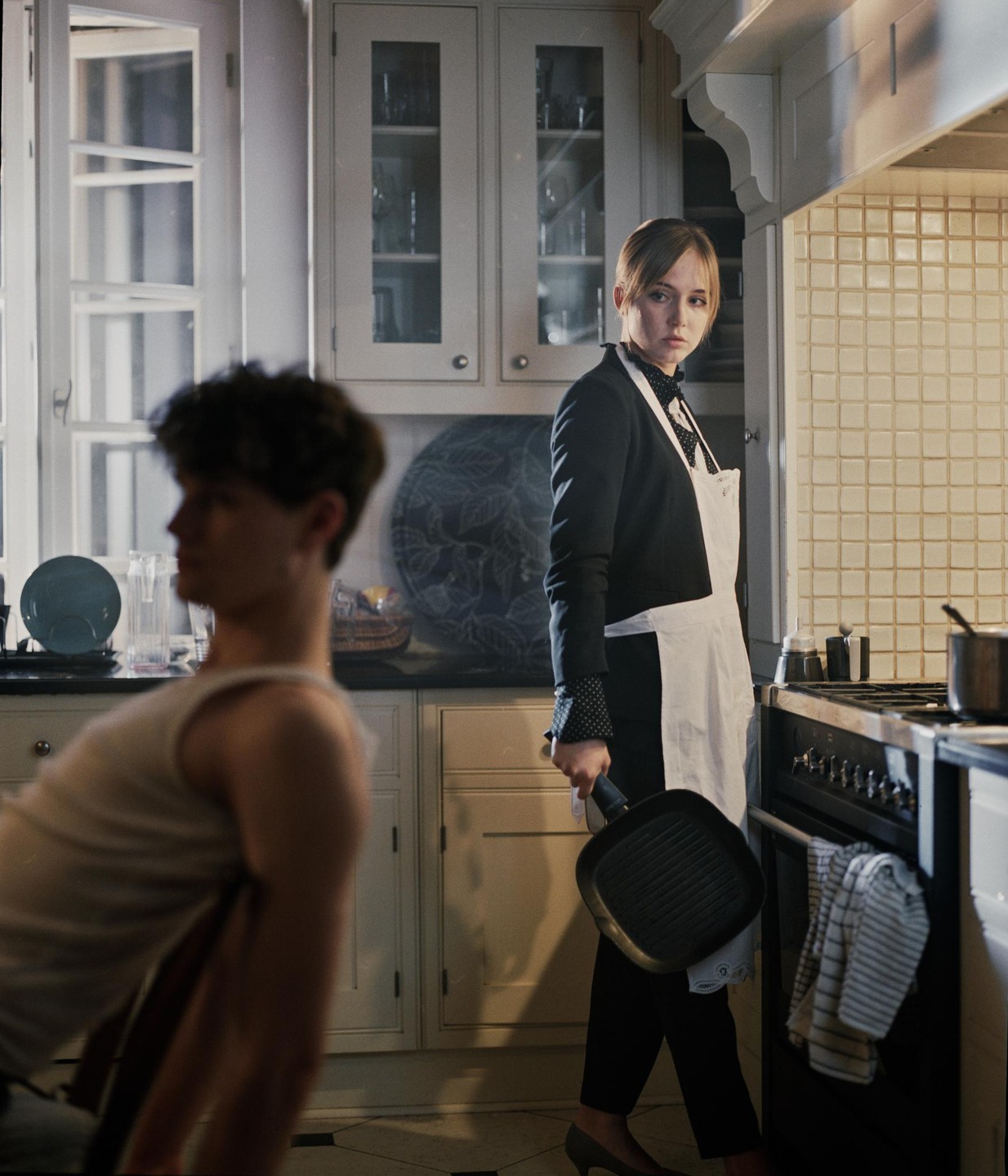 A woman in an apron holds a frying pan in a kitchen while a man sits in the foreground.