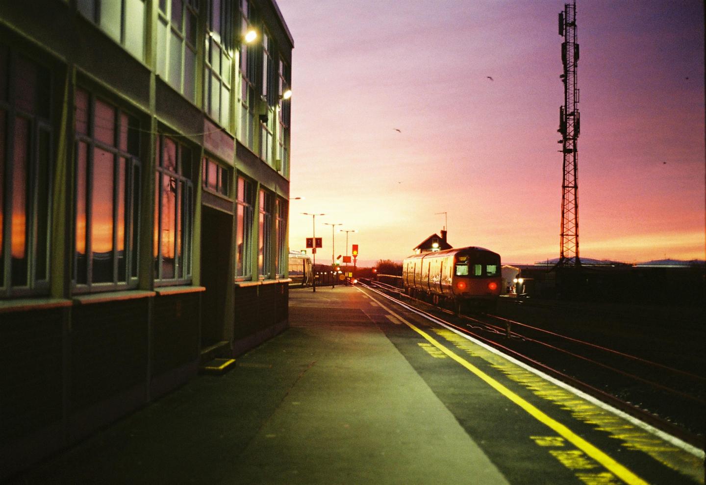 A train is stopped at a station platform at sunset, with a building on the left and a large antenna tower on the right.