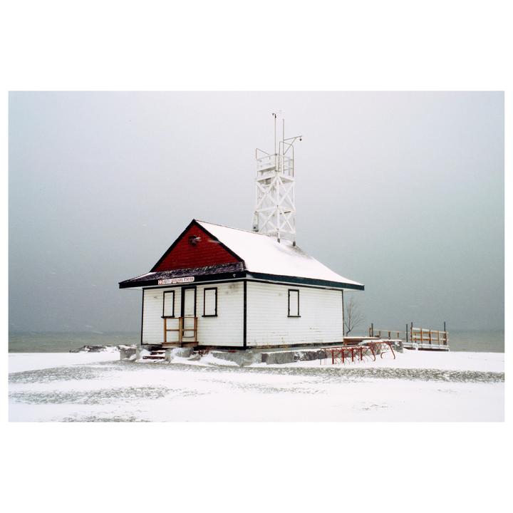 A small white building with a red roof and a tall white tower stands in a snow-covered landscape next to a body of water.