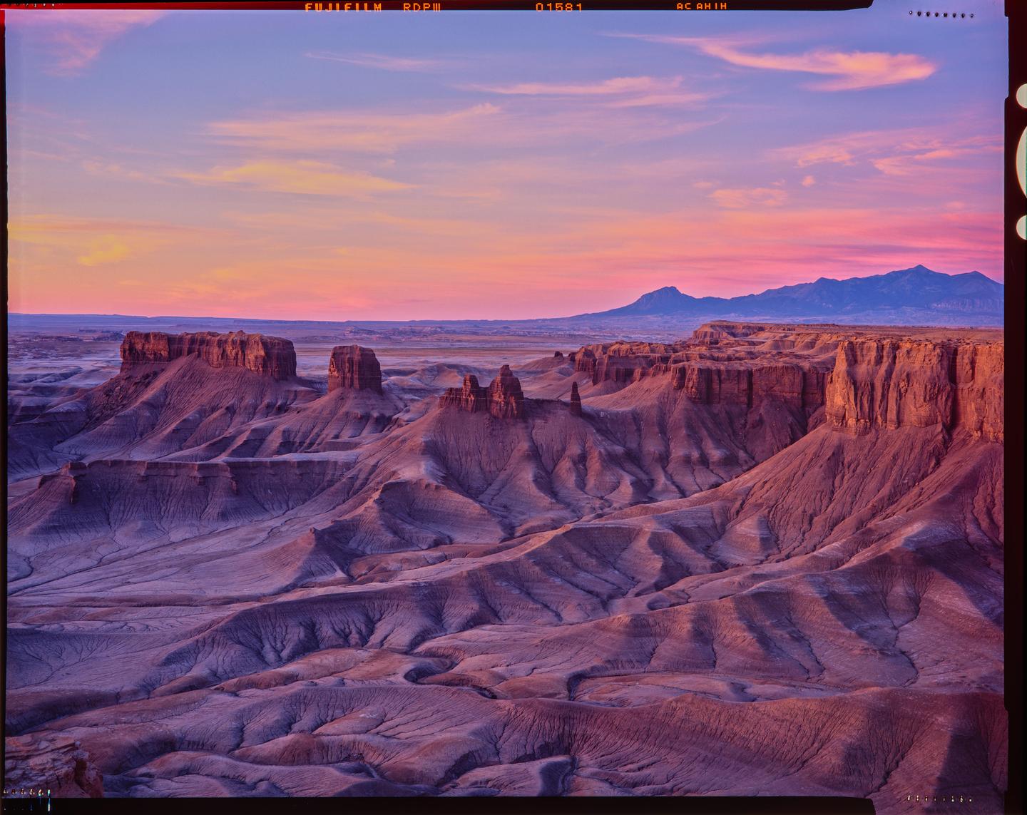 A vast desert landscape with mesas and mountains is illuminated by the colorful light of sunset.