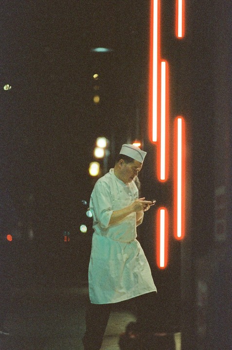 A man in a chef's uniform looks at his phone on a dark street illuminated by red neon lights.
