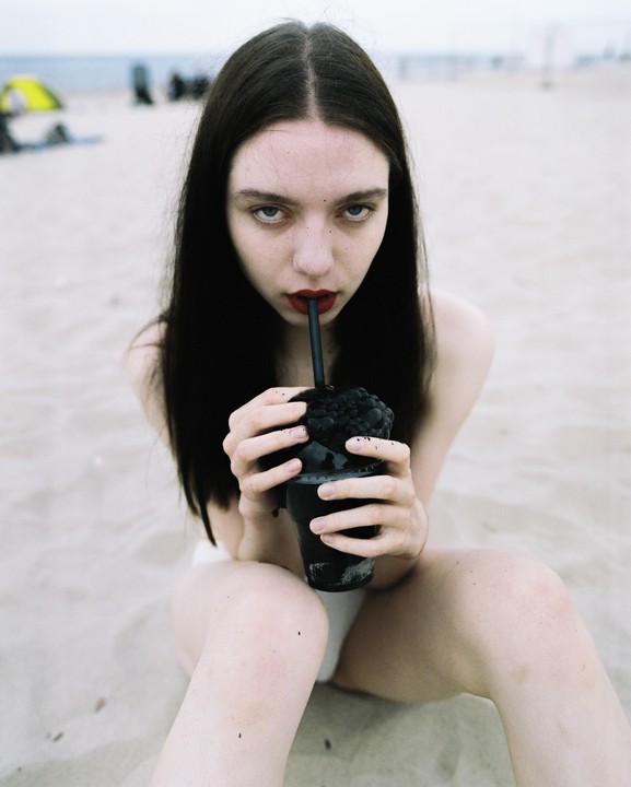 A woman with long dark hair and red lipstick sits on a sandy beach, holding a black cup with a straw.