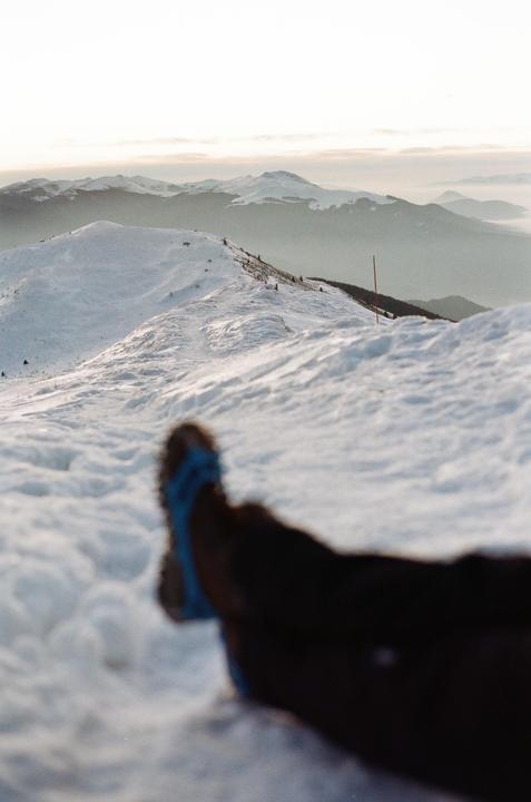 A person's legs and feet are visible in the foreground of a snowy mountain landscape.