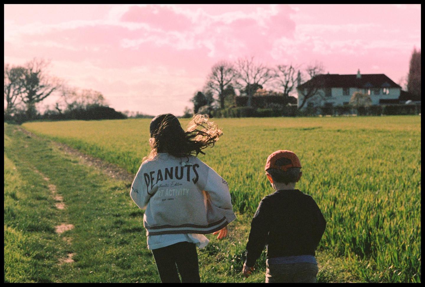 Two children walk away from the camera through a field of green grass toward a house under a pink sky.