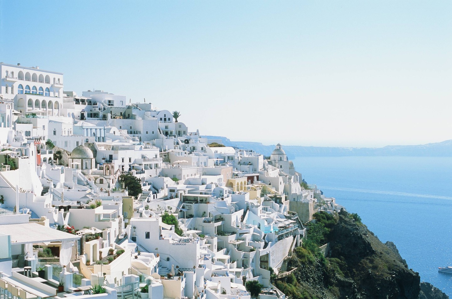 A white village with many buildings and a church with a dome is built into a cliff overlooking a calm blue sea.