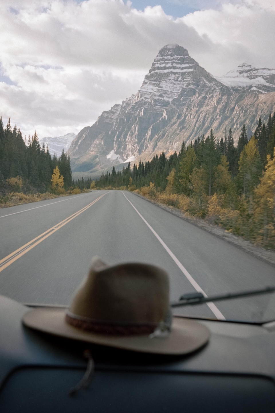 A hat sits on a dashboard, looking out at a road leading to a snow-capped mountain surrounded by forest.
