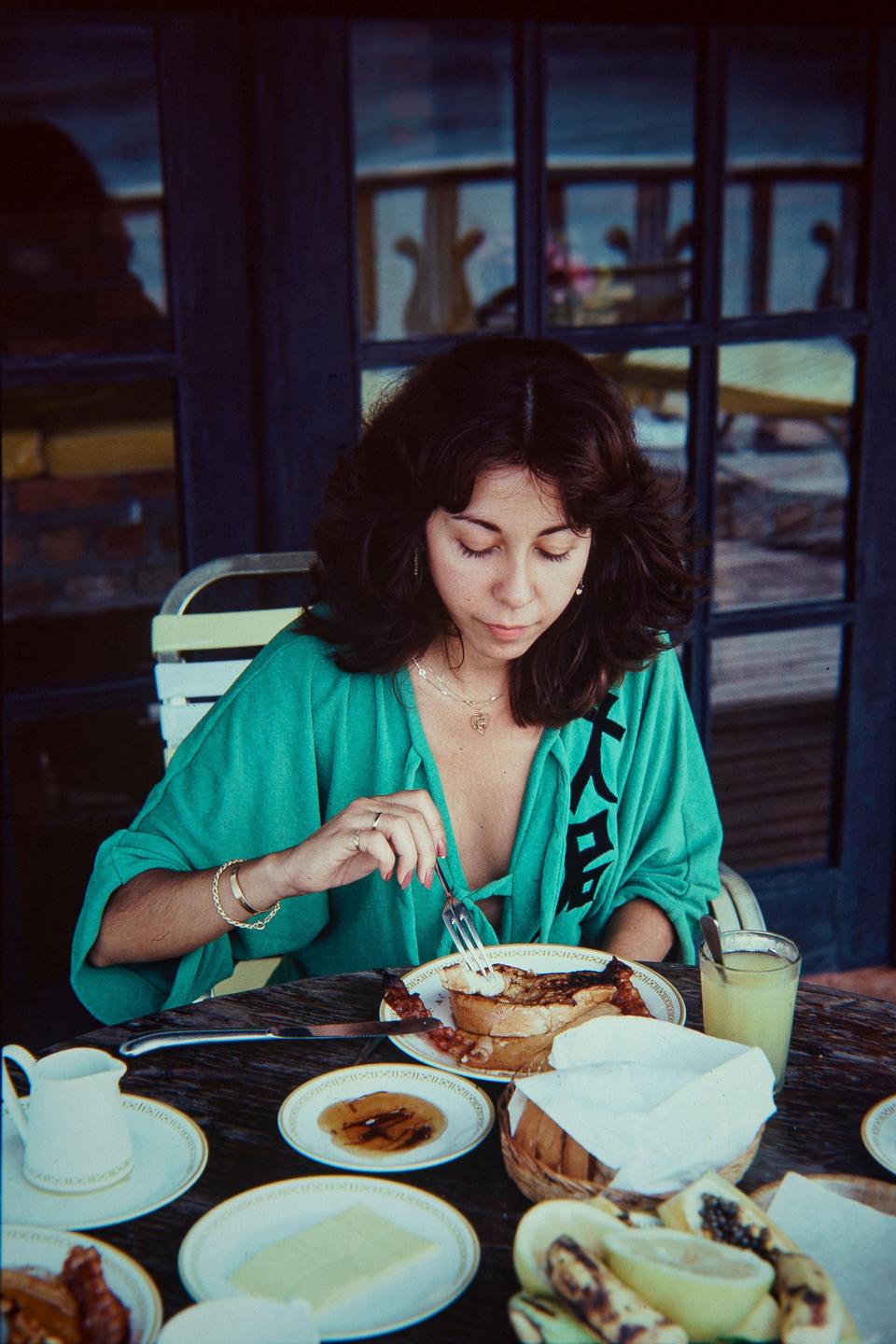 A woman in a green shirt eats breakfast at a table laden with food and drink.