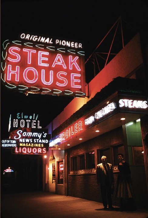 A couple stands outside a steak house and hotel with neon signs at night.