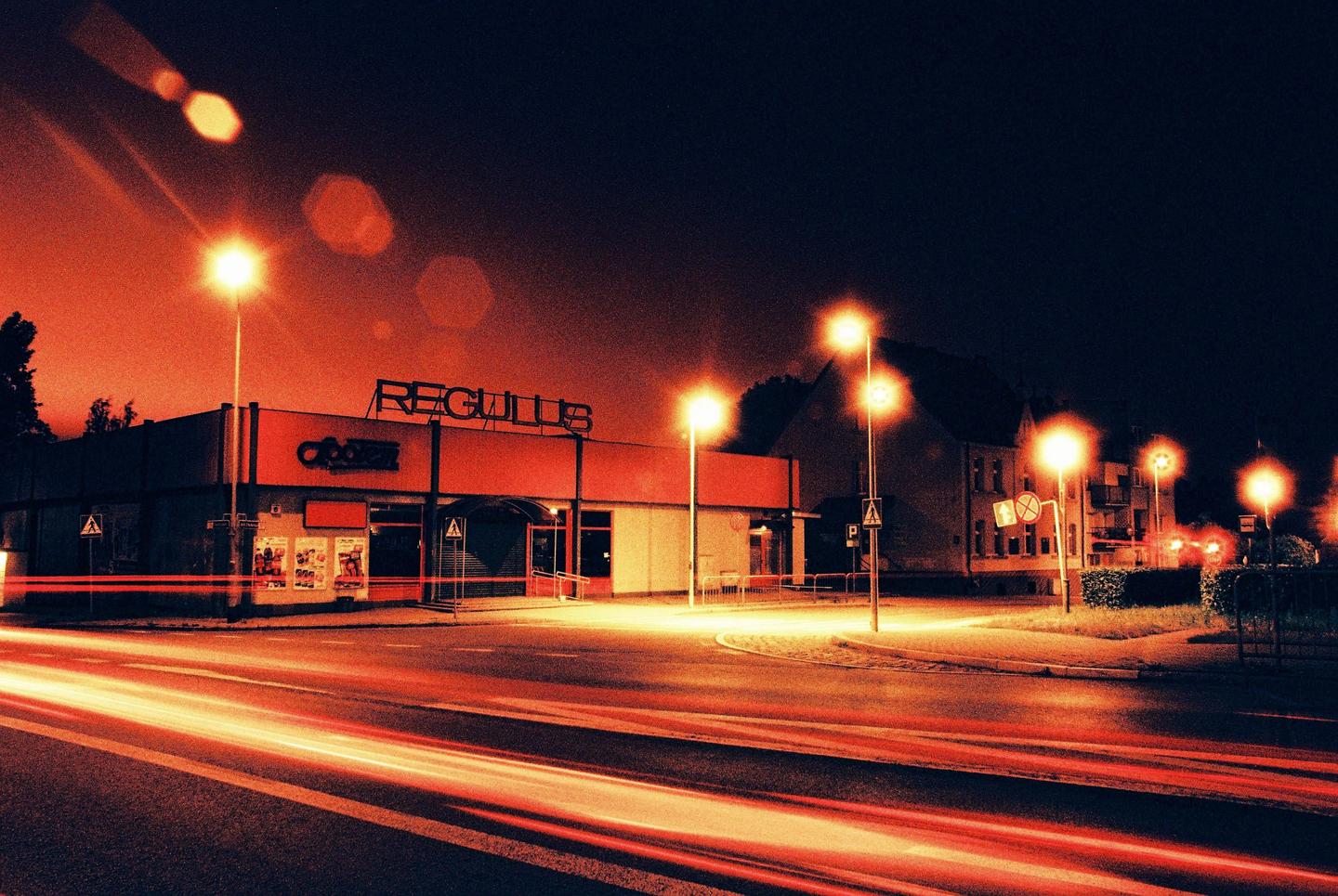 A street at night shows a building with the name "REGULUS" on it, with light trails from vehicles on the road.