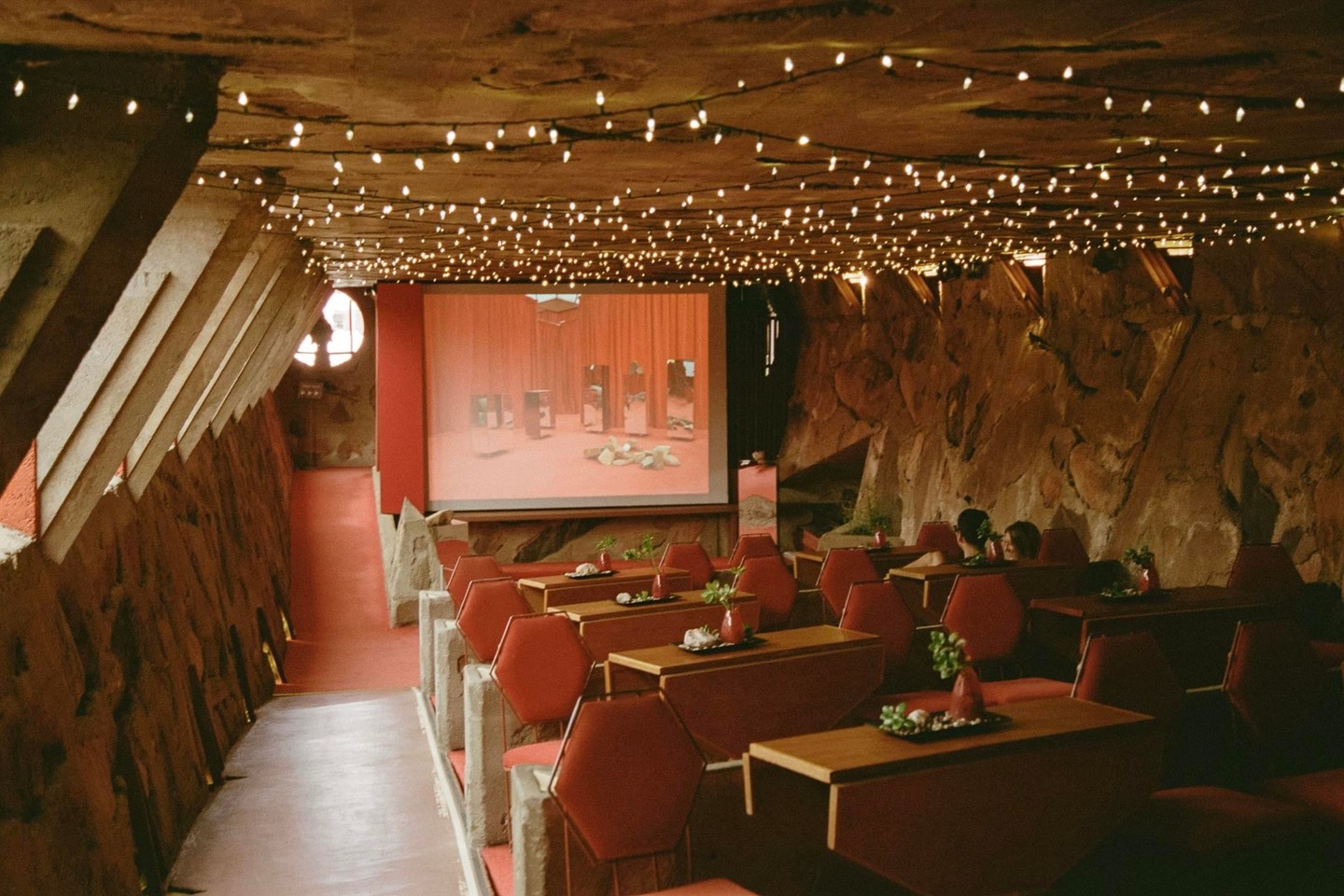 A movie theater with red seats and tables is illuminated by string lights on the ceiling and a film projected on the screen.