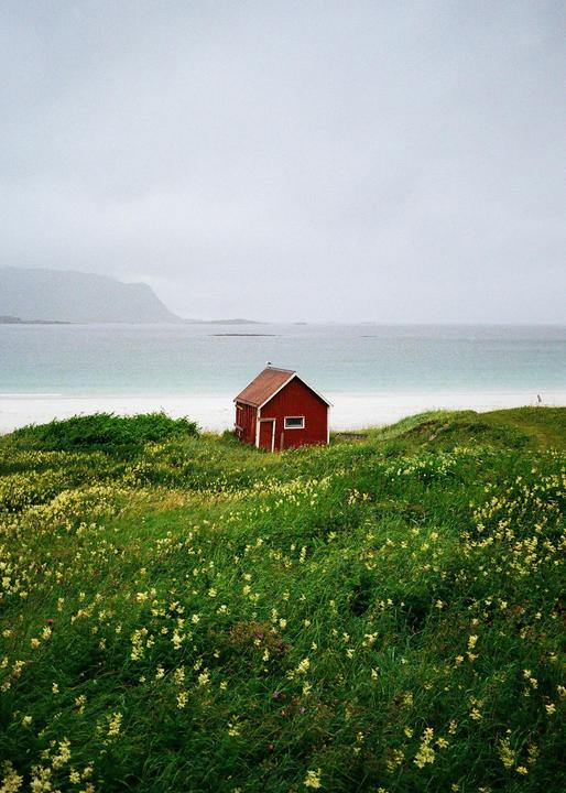 A red cabin sits on a grassy hill overlooking a white sand beach and the ocean under a cloudy sky.