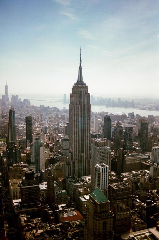 The Empire State Building rises above the surrounding cityscape with a body of water and distant land visible in the background.