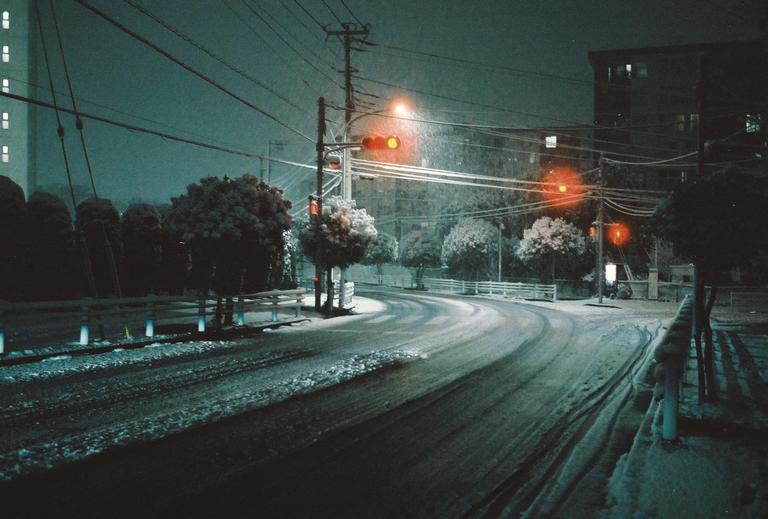 A snow-covered street curves past trees and buildings under a dark night sky with glowing traffic lights.