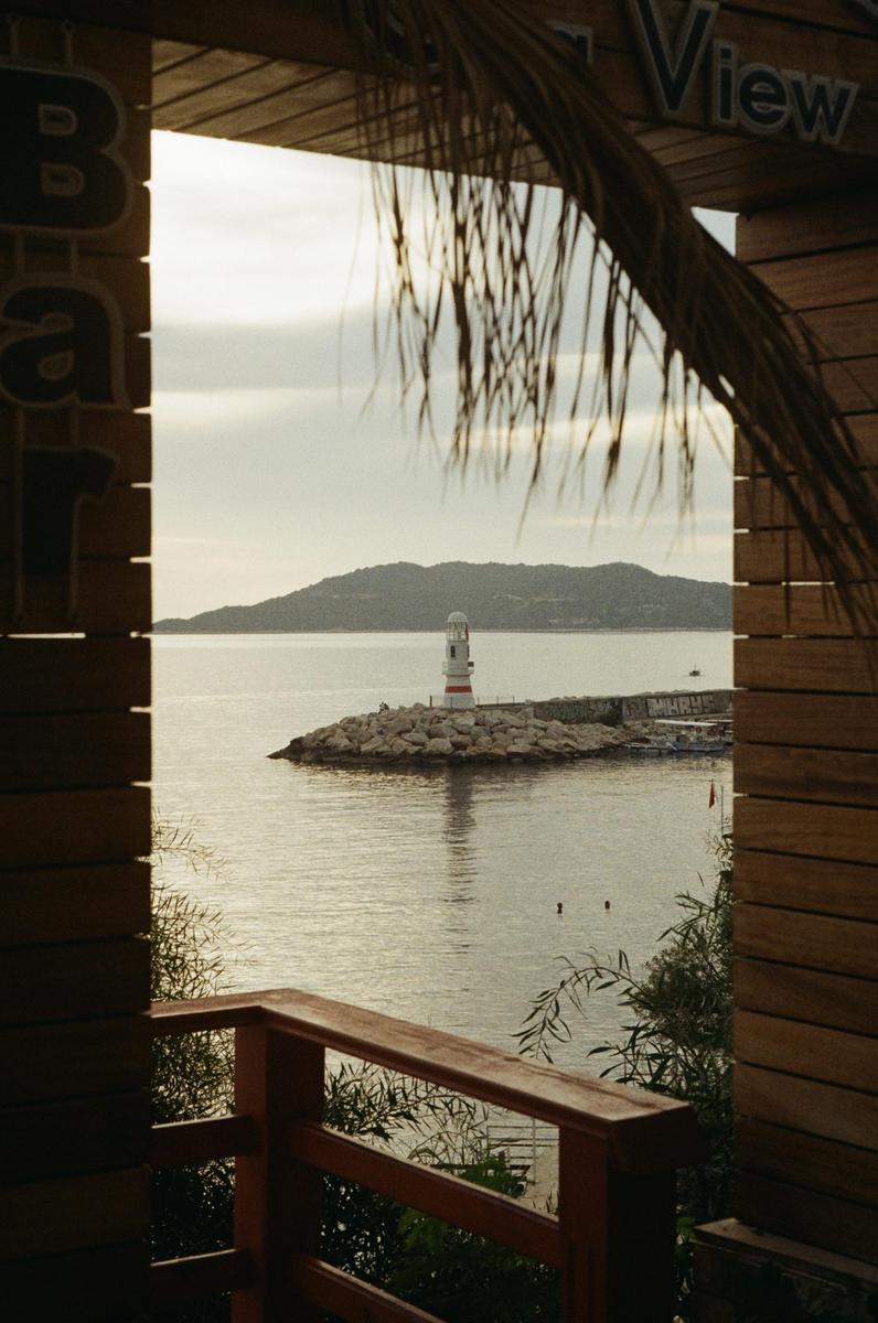 A lighthouse stands on a rocky pier in the middle of a body of water, framed by a wooden structure and palm fronds.