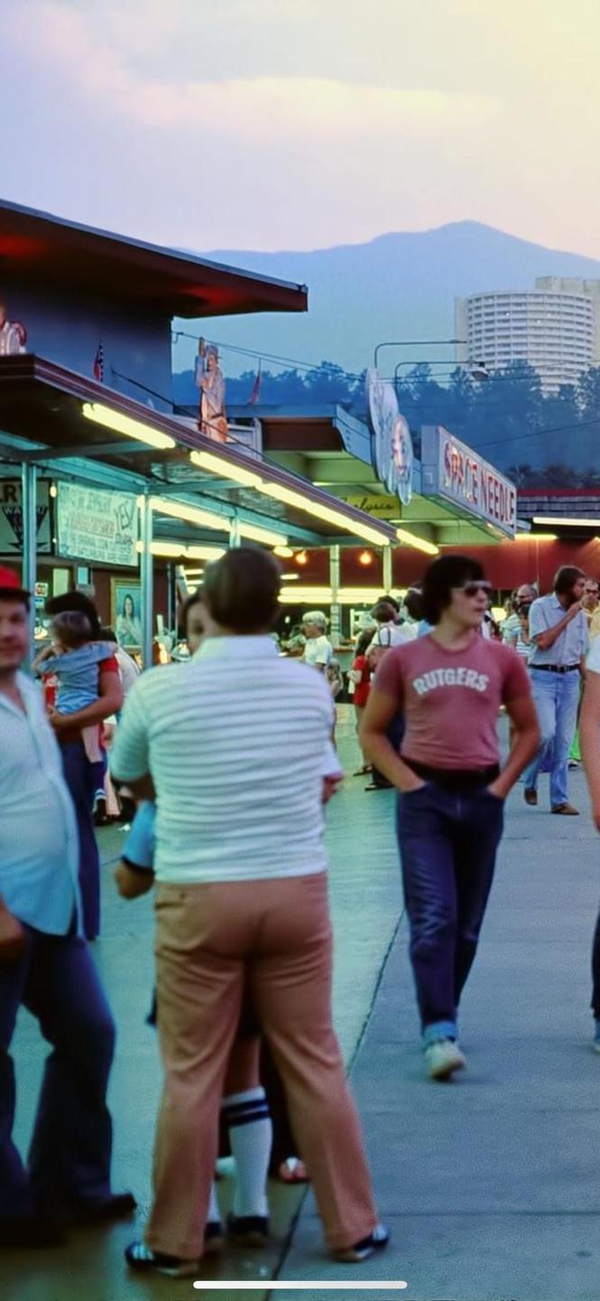 A crowd of people walk along a street with buildings and mountains in the background.