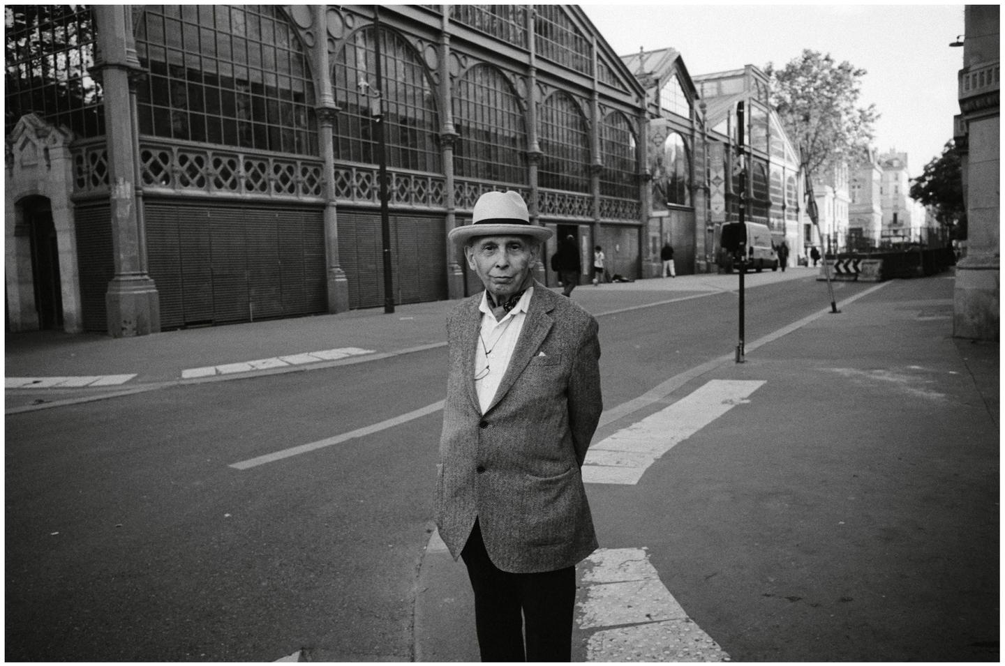 A man in a hat and jacket stands on a street in front of a large building with arched windows.
