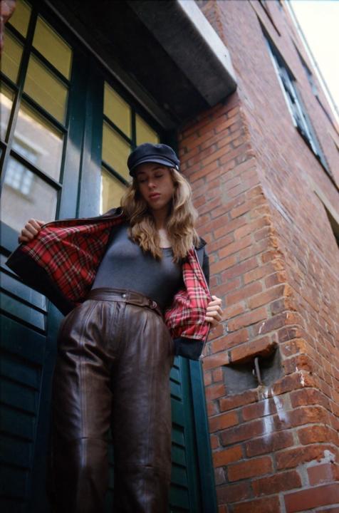 A woman wearing a cap and a jacket with a plaid lining stands in front of a brick building.