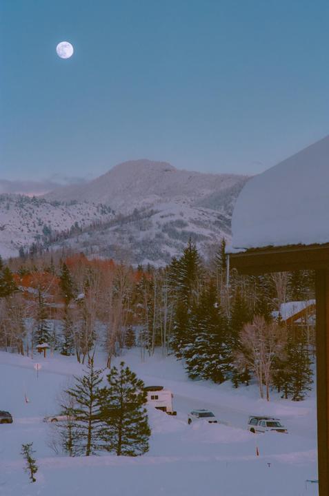 A full moon hangs in a blue sky over a snow-covered mountain landscape with trees and parked vehicles.
