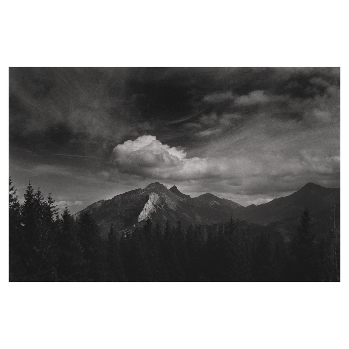 A dramatic black and white landscape shows a mountain range with a single cloud above the peak, and a forest in the foreground.