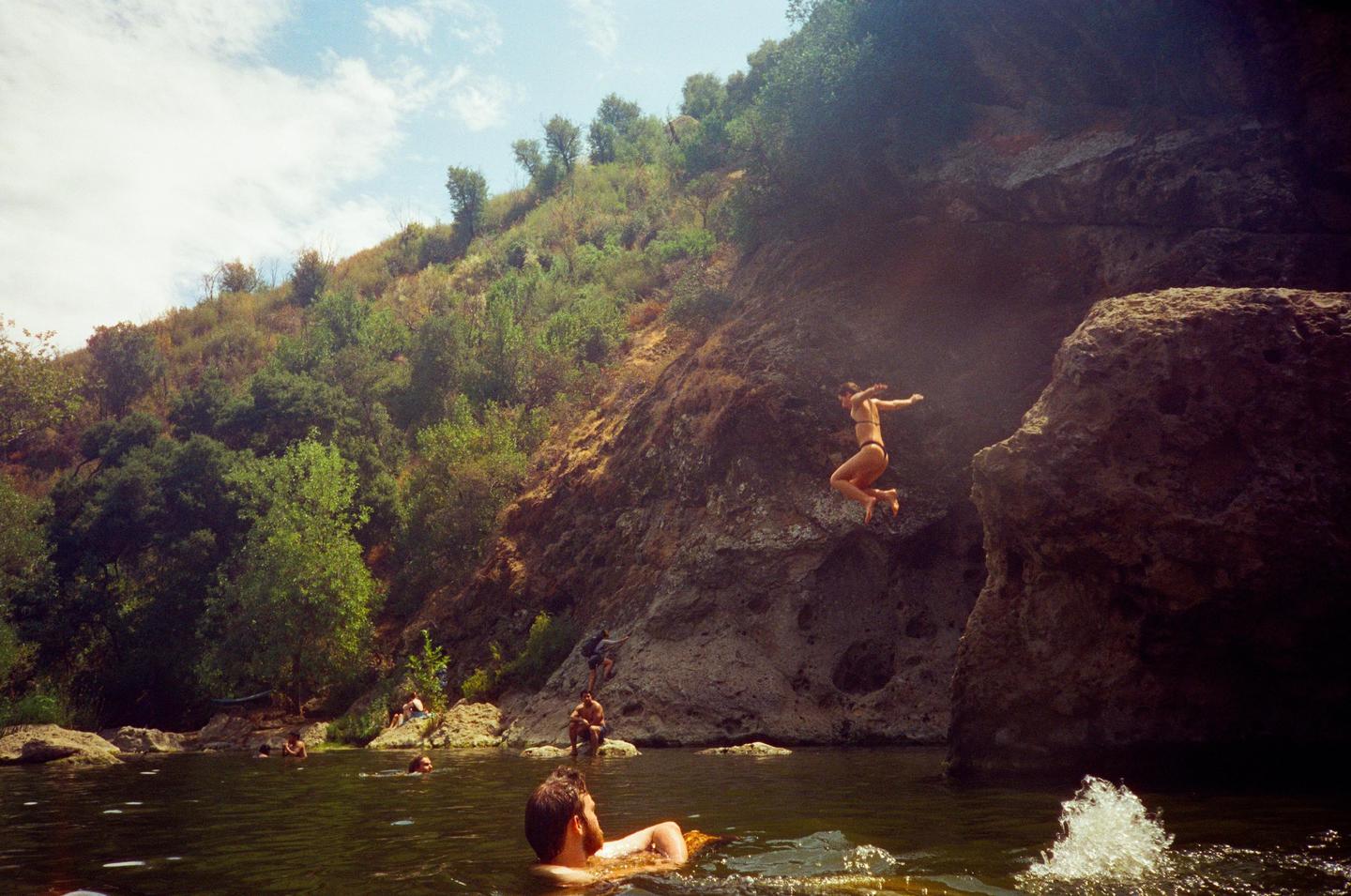 A woman jumps from a rock into a swimming hole with other people in the water and on the shore.