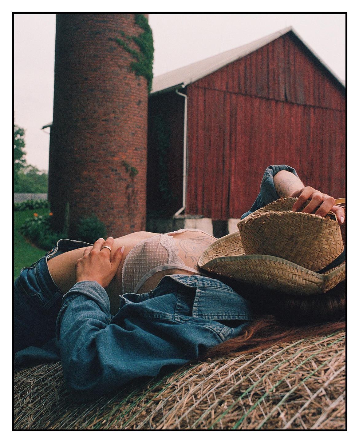 A woman wearing a cowboy hat and denim jacket is lying on a hay bale in front of a red barn and brick silo.